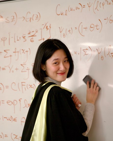 Ziyi (Ellen) Zhou in a graduation gown, wiping a whiteboard covered in copula formulas